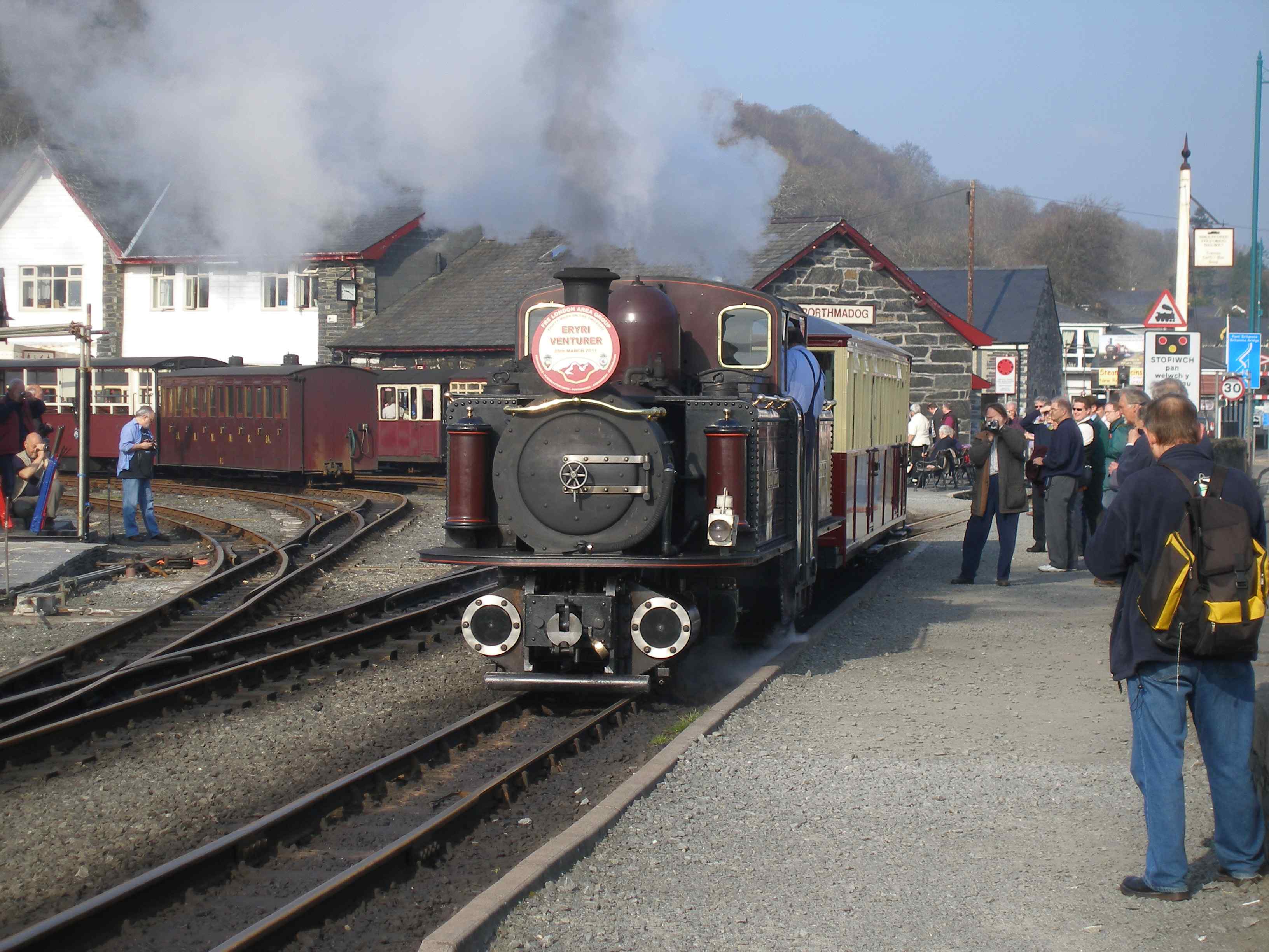 Ready for the off. 'Merddin Emrys' stands at Porthmadog pointing towards Blaenau Ffestioniog. The loco is standing on the connection to the Welsh Highland Railway, which passes to the right of the old goods shed in the background, and then becomes a street tramway as it crosses Britannia Bridge, before swinging north onto its dedicated alignment. Behind 'Merddin' is car 123 aka 101. This was an original first class Observation Car but was to be replaced. However on being assessed it was found to be in fairly good condition, so was given a new lease of life as a Third Class Observation Car. Although the internal paintwork was not quite finished, this was its first day in traffic. marshalled at the top end of the train (FR Observation Cars are traditionally at the bottom or downhill end of trains).