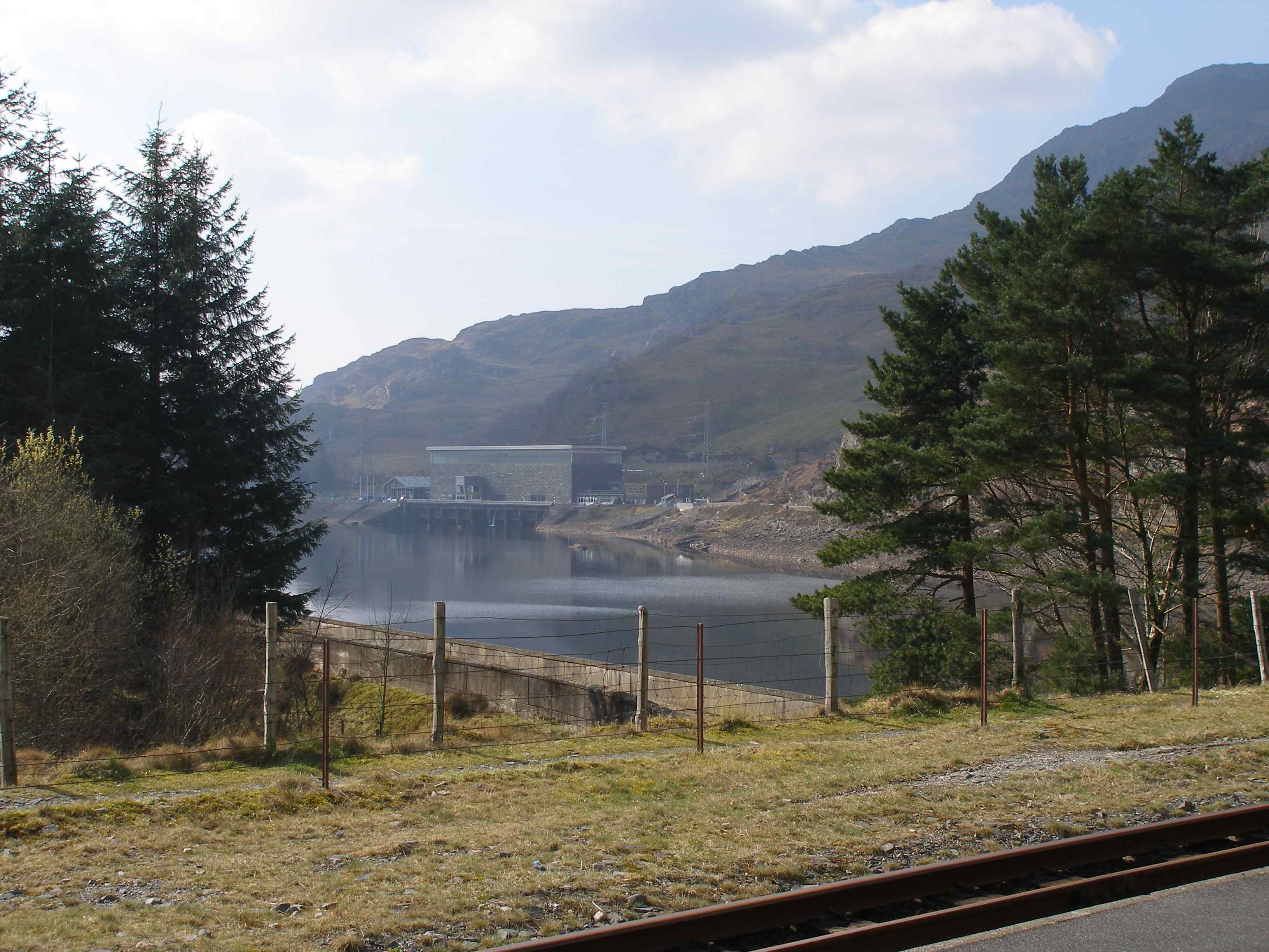 The pumped-hydro electric power station from Tan-y-Grisiau station. The line used to run along the road leading to the power station and then through the actual site - the old cutting on the other side is still visible. The new line now climbs from the station before emerging just below the roof line of the Power Station, level with the upper windows. The ballast bed can just be made out. Commissioned in 1963, Ffestiniog Power Station was the UK's first major pumped storage power facility. Although of an older generation to those at Dinorwig, Ffestiniog's four generating units are still capable of achieving a combined output of 360MW of electricity - enough to supply the entire power needs of North Wales for several hours. The Generation Cycle begins at Llyn Stwlan - Ffestiniog's upper reservoir. Large screens inside the intake towers are opened to activate the high-pressure downflow. 